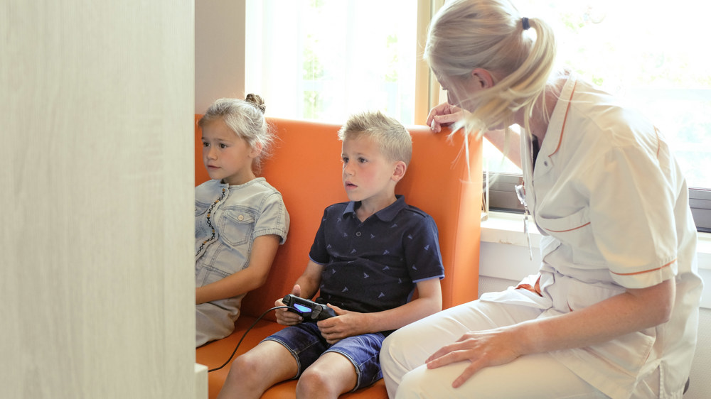 2 kinderen gamen in de speelkamer en de verpleegkundige kijkt mee. 2 kinderen gamen in de speelkamer en de verpleegkundige kijkt mee.