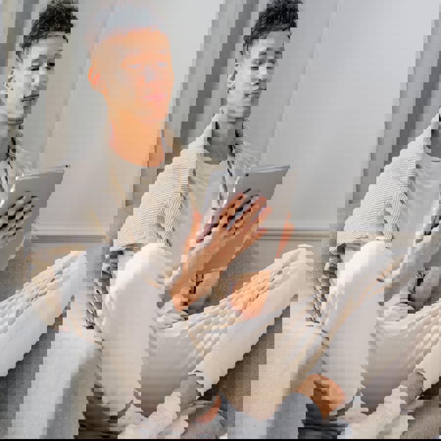 Jongeman die in kleermakerszit in een stoel op een tablet zit te kijken Jongeman die in kleermakerszit in een stoel op een tablet zit te kijken