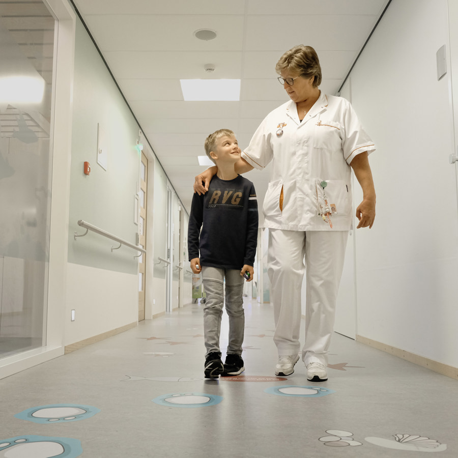 Links een kind, rechts een verpleegkundige. De verpleegkundige slaat een arm om het kind. Ze lopen in een gang. Links een kind, rechts een verpleegkundige. De verpleegkundige slaat een arm om het kind. Ze lopen in een gang.
